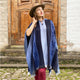 A person wearing a blue poncho and beige hat stands on cobblestone steps. They hold the hat's brim, with a wooden door and stone building in the background.
