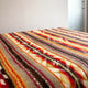 Colorful patterned blanket lies on a bed, featuring geometric designs in red, brown, and beige. The setting includes a minimalist room with a gray wall and glass door in the background.