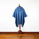 A blue poncho with a hood drapes over a wooden mannequin stand, set against a white wall and hardwood floor.