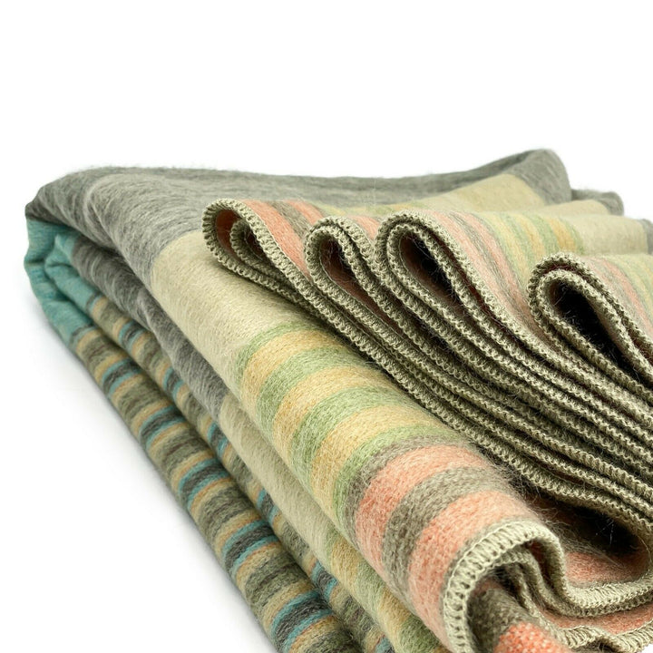 Cojitambo alpaca wool throw blanket – folded woolen blankets display alternating pastel stripes of green, peach, and beige. They are layered on top of each other, suggesting a cozy, neatly arranged setting