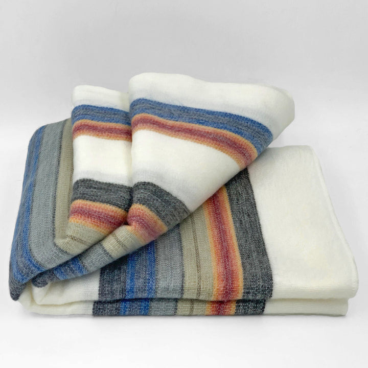 Siguitag alpaca wool throw blanket – a folded blanket features broad horizontal stripes in blue, orange, and gray against a soft white background. It rests neatly arranged in a plain setting
