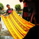 A person holds a vibrant yellow, patterned textile, displaying it outdoors in a marketplace. They wear a gray shirt with the text Yellow Time in pink.