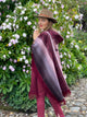 A person wearing a dark red poncho and brown hat stands among lush green bushes with light purple flowers on a stone pathway.