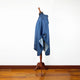 A blue hooded poncho is draped on a wooden mannequin stand, positioned in a room with a wooden floor and white wall.