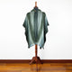 A dark green poncho with intricate patterns is displayed on a wooden mannequin stand against a white wall, set on a polished hardwood floor.
