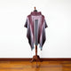 A purple and gray poncho with fringe hangs on a wooden mannequin stand, set against a plain white wall and wooden floor.
