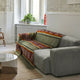 A gray sofa is covered with a multicolored patterned blanket in a cozy living room. It is positioned near a window with potted plants on a white radiator and light-colored walls.