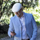 A man in a light blue suit and white flat cap is looking at his phone while walking outdoors, surrounded by greenery.