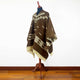 Clint Eastwood alpaca wool poncho – a brown poncho with white geometric patterns drapes over a mannequin, standing on a wooden floor against a plain white wall. There is no text in the image