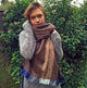 A person wearing a large, brown scarf and gray sweater stands in a garden setting with lush green foliage and small pink flowers in the background.