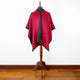 A red cape with a black trim is displayed on a wooden mannequin stand, set against a plain white wall with a wooden floor.