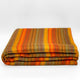 Cumanda baby alpaca wool blanket – a folded, multicolored striped blanket with shades of orange, yellow, brown, and green, placed against a plain white background