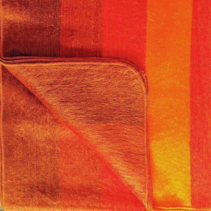 Yacupungu baby alpaca wool blanket – a colorful blanket displays vivid orange and red horizontal stripes, lying flat with one corner folded over, revealing its texture and vibrant hues in bright lighting