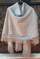 A gray shawl drapes elegantly on a hanger, showcasing intricate crochet edging and long fringes, set against a decorative wrought iron backdrop.