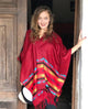A person wearing a red, multi-patterned poncho stands smiling in a wooden doorway, holding a hat. The setting appears to be a dimly lit interior space.