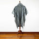 A gray poncho with intricate geometric patterns drapes over a wooden stand, set against a plain white wall and wooden floor.