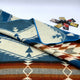 A folded blanket with geometric patterns lies on a surface, showcasing blue, brown, and cream colors. Nearby, small sachets of various spices or condiments are scattered.