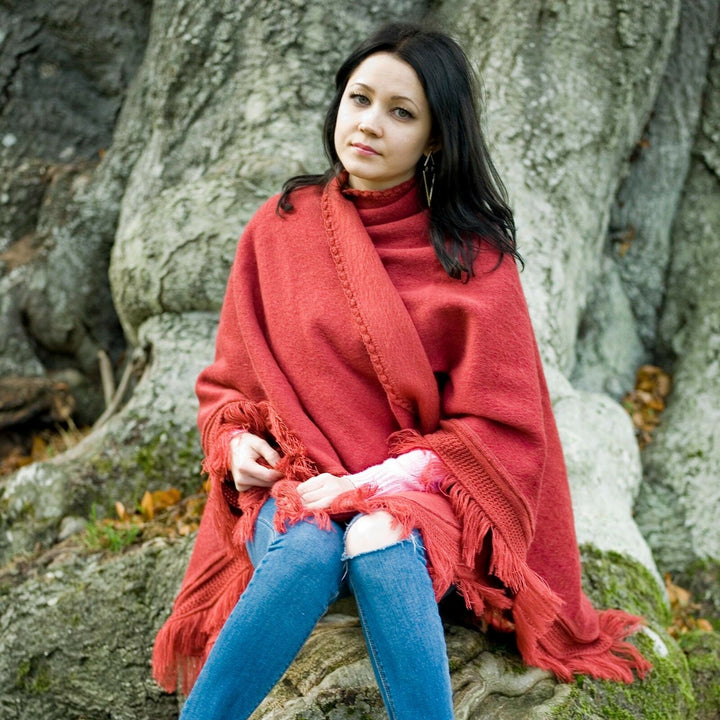 A person wearing a red shawl sits against a large tree trunk, surrounded by textured bark and scattered leaves. The person appears relaxed in a natural outdoor setting.