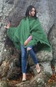A person wearing a green shawl and ripped jeans stands against a large tree trunk. Autumn leaves cover the ground, creating a natural, rustic scene.