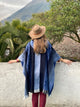 A person wearing a hat and blue shawl stands facing away, leaning on a white wall, surrounded by trees, with a mountain visible in the background.