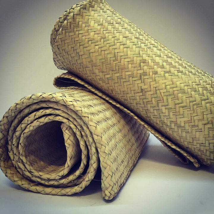 Two rolled straw mats lie on a surface. One mat is partially unrolled, displaying a woven pattern. The mats are in a neutral setting with soft lighting.