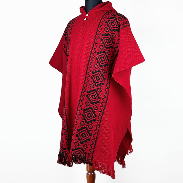 A red poncho with black geometric patterns runs vertically, displayed on a mannequin against a plain white background. The poncho features fringed edges at the bottom.