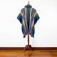 A colorful poncho with geometric patterns drapes over a mannequin, set against a white wall backdrop and wooden floor.