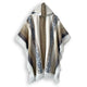 A hooded poncho featuring geometric patterns and fringed edges hangs against a plain white background. The colors blend browns, whites, and grays, creating an earthy, woven texture.