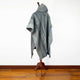 A gray, patterned poncho drapes over a wooden mannequin stand, displayed in a room with wooden flooring against a plain white wall.