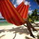 A red and orange hammock holds a person relaxing, with their legs visible. It hangs between palm trees on a sandy beach, overlooking a turquoise ocean under a blue sky.