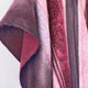 A draped woolen fabric, featuring warm tones of burgundy and pink, hangs against a plain white wall. The texture is soft, with subtle patterns and gradients blending throughout.