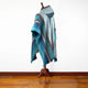 A multicolored poncho with blue and grey stripes drapes over a wooden mannequin stand, set against a plain white wall and wooden floor.