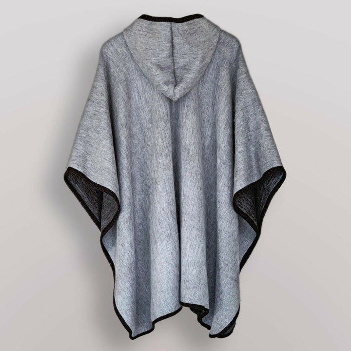 A gray hooded poncho hangs against a plain wall, showcasing its loose, flowing fabric and black-trimmed edges.