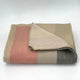 Tilivi baby alpaca wool blanket – folded blanket lying on a flat surface, displaying striped patterns in muted tones of beige, pink, and gray. The background is a plain, light-colored surface, providing a simple setting