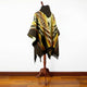 A colorful poncho with geometric patterns drapes over a wooden mannequin in a room with light-colored walls and a polished hardwood floor.