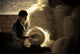 A person inspects a straw hat, surrounded by large bundles of straw. Soft light filters through a window, highlighting the intricate texture of the straw and the brick wall.