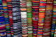 Colorful woven textiles hang vertically, displaying vibrant patterns and stripes, within a market setting.