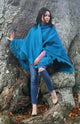 A person wearing a blue fringed shawl and ripped jeans leans against a large, textured tree trunk, surrounded by autumn leaves.