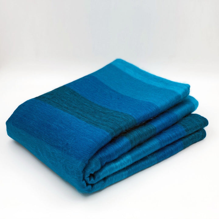 Cajabamba alpaca wool blanket – a folded blue blanket displays varying shades, resting neatly. The background is plain and light, enhancing the vibrant colors of the fabric