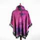 A vibrant magenta poncho, featuring black tie-dye patterns, drapes elegantly on a wooden hanger. The garment has fringed edges and is set against a plain white background.