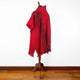 A red poncho with intricate black geometric patterns draped over a wooden mannequin stand, set against a plain white wall and wooden floor.