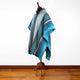 A multicolored poncho with blue and gray stripes displayed on a wooden mannequin stand in a room with a wooden floor and a plain white wall.