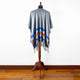 A gray poncho with colorful patterns and fringe drapes over a mannequin. It stands on a wooden floor against a plain white wall, showcasing its design.
