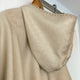 A cream-colored woolen poncho with a hood hangs on a wooden hanger against a white wall, showcasing its soft texture and detailed stitching along the edges.