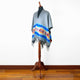 Gray poncho with colorful geometric designs and fringe displayed on a wooden mannequin stand. It stands against a plain white wall on a polished wooden floor.