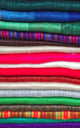 Colorful woolen fabrics are stacked in layers, showcasing diverse patterns and vibrant hues, including green, blue, red, and striped designs, creating a visually rich and textured display.