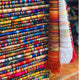 Stacks of brightly colored, woven textiles with intricate patterns are folded neatly, showcasing vibrant geometric designs. Two rolled-up fabrics with fringed edges hang to the side.
