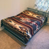 A bed with a metal frame has a patterned blanket featuring geometric designs. It is placed in a carpeted room by a wall with floral curtains partially visible.