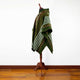A green poncho with white and brown stripes is displayed on a wooden mannequin stand against a plain white wall, resting on a polished wooden floor.