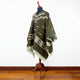 Clint Eastwood alpaca wool poncho – a green poncho with white geometric patterns is displayed on a mannequin stand. The poncho features fringed edges and is set against a wooden floor and white background